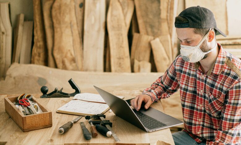 A craftsman wearing a mask, working on a laptop in a woodworking shop.