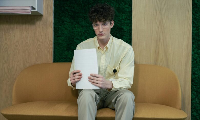 Young man in yellow shirt and glasses sits on sofa in an office, holding papers.