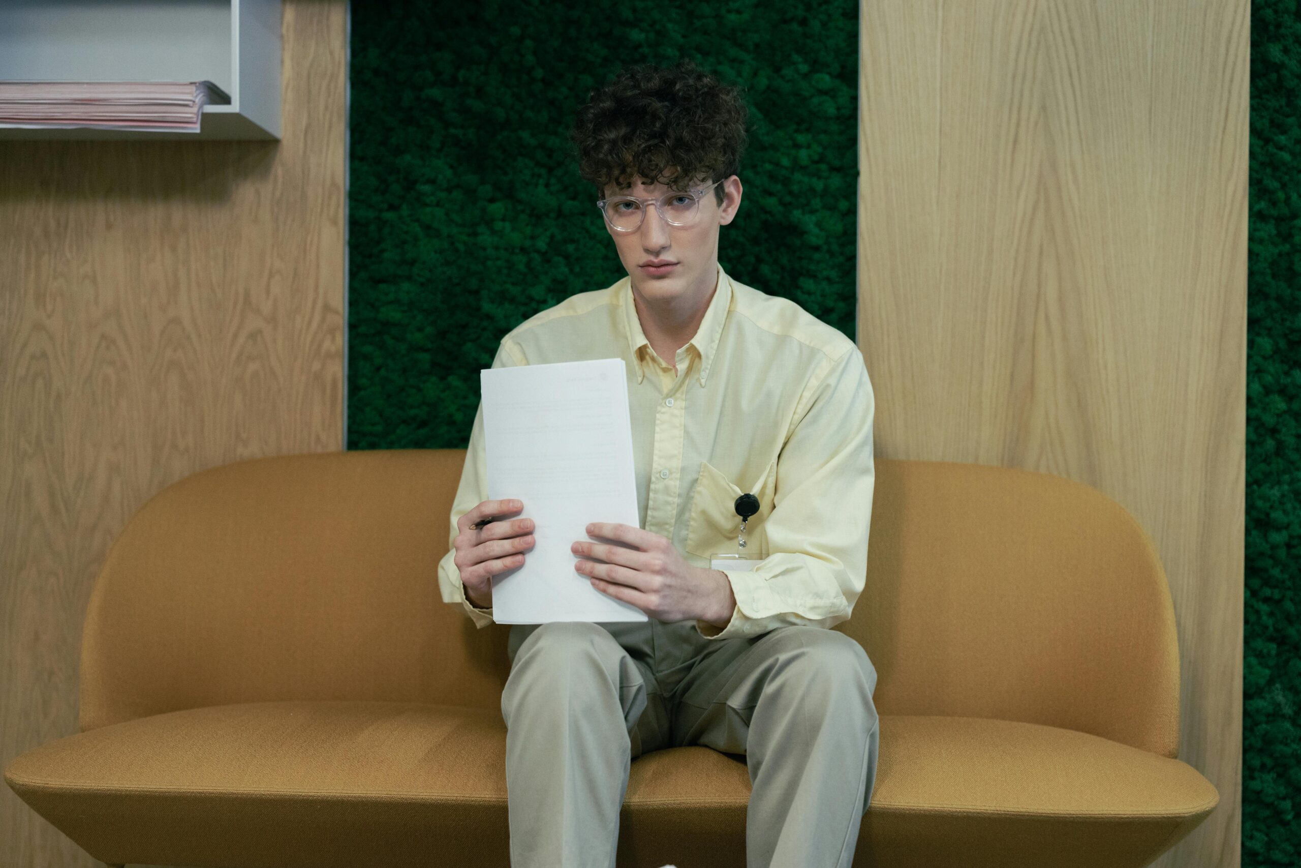 Young man in yellow shirt and glasses sits on sofa in an office, holding papers.